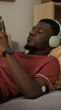 Vertical of young adult Black man with wearable glucose sensor listening to audio content through headphones while relaxing on sofa in living room
