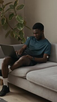 Vertical full length shot of young adult Black man wearing CGM sensor typing on laptop while relaxing on sofa in plant decorated modern living room, slow motion