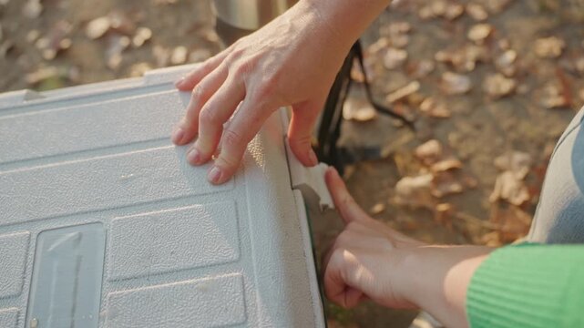 Hands opening camping cooler on leaf strewn ground, textured plastic lid and latch under warm autumn sunlight, camper preparing chilled drinks, sneakers and backpack visible, close up detail showing