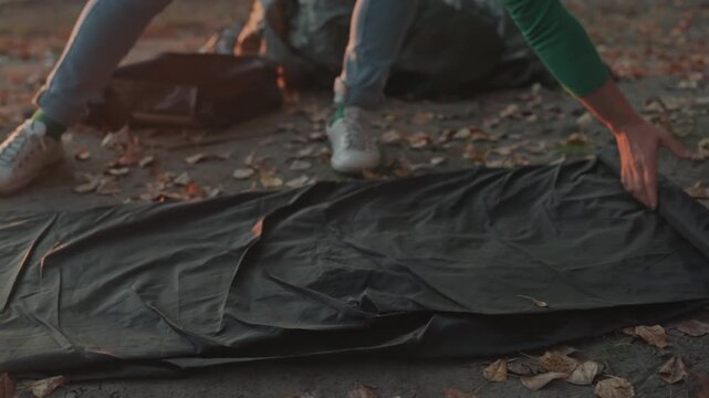 White camper unrolling sleeping bag on pavement strewn with autumn leaves, hands smoothing fabric and straightening seams, jeans and sneakers visible in warm dusk light preparing bivouac at campsite,
