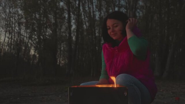 Woman warming hands at campfire dusk in forest clearing, gentle embers glow, cozy pink vest, kneeling by metal fire pit, stoking small flame, reflective expression, soft evening light, peaceful