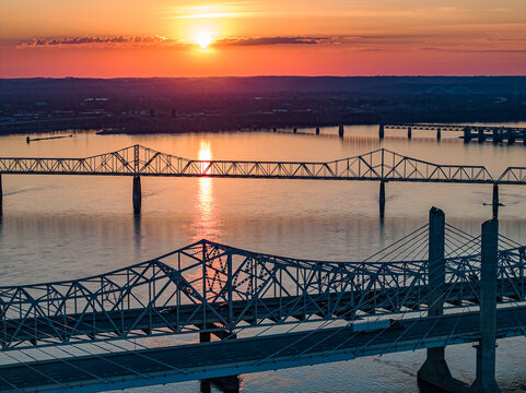 Aerial view of multiple bridges spanning the Ohio River during a vibrant sunset with orange sky reflections in Louisville, Kentucky, United States.