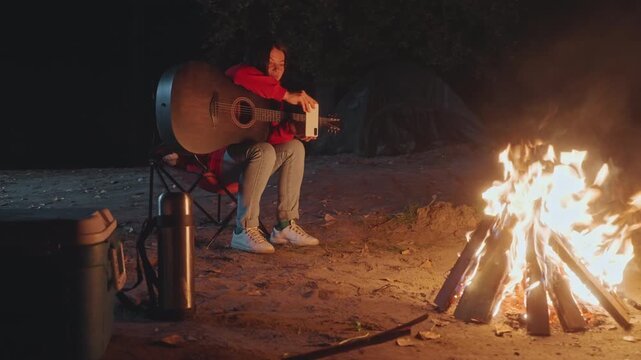 Calm campfire guitar moment asian male leaning on acoustic instrument beside crackling bonfire, warm glow on red jacket, quiet reflection, camping night solitude, soft ember light and rustic ground
