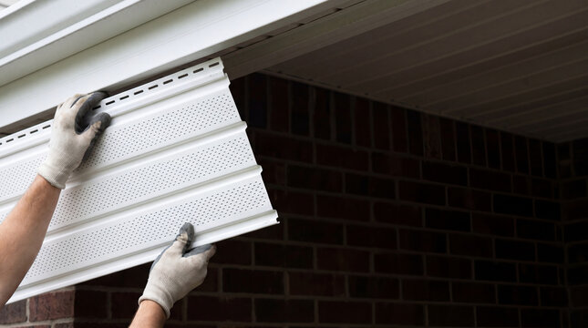 Roofer installing white vinyl soffit panel under roof eave. Construction worker repairing house exterior. Building renovation, home improvement and maintenance project concept.