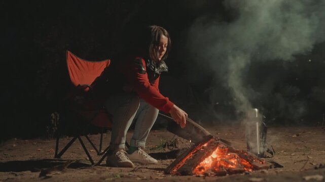 Black woman stoking campfire creating spark shower, throwing log into embers with deliberate motion, smoke plume and lantern glow highlight intense camping moment in night wilderness