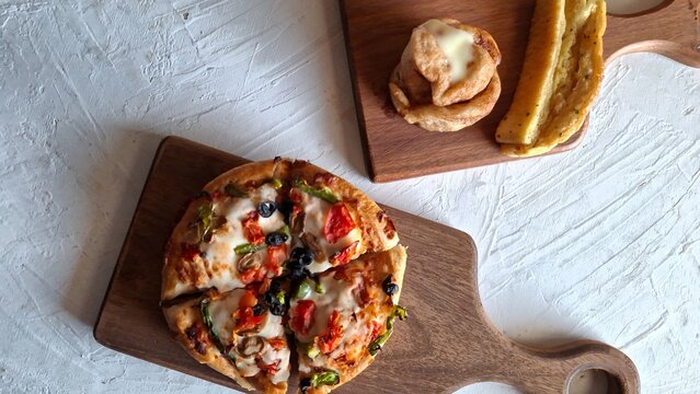 Small Homemade Veggie Pan-Style Pizza Served on Rustic Wooden Cutting Board with Cinnamon Roll and Slice of Breadstick on Textured White Surface
