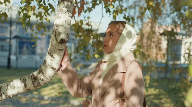 Caucasian woman headscarf touching birch, sunlit park autumn leaves, gentle shadow patterns on coat, contemplative gaze, slow hand movement along bark scenes alternate between quiet stroll, memory