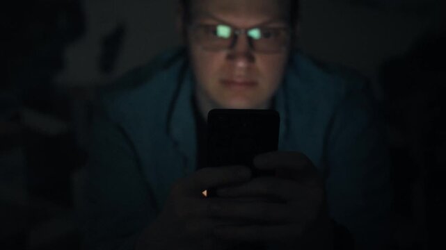 In dim bedroom at night, bespectacled man grips his smartphone, staring at bright screen. His late-night scrolling shows harmful dependence that disrupts healthy sleep, highlighting tech intrusion