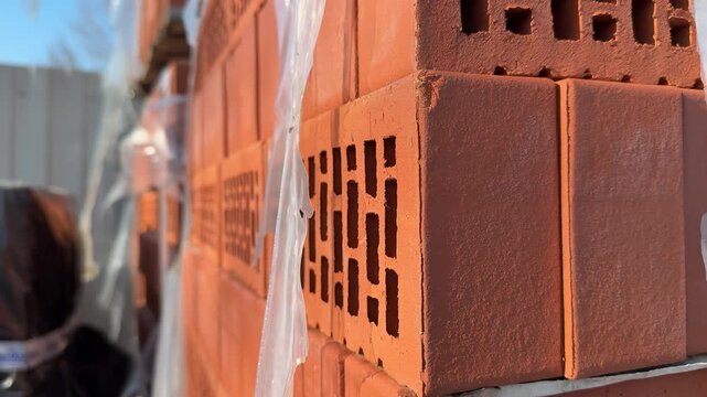 Close-up red bricks stacked outdoors, plastic wrap glistening in sunlight, perforated hollow bricks on pallet at hardware store