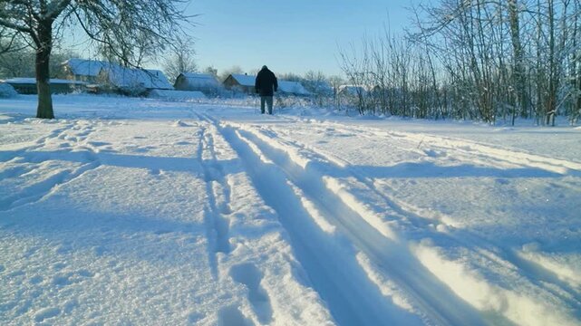 A man in winter clothing skis across a snowy field, leaving clear footprints in the snow, while trees and distant houses frame a serene winter landscape
