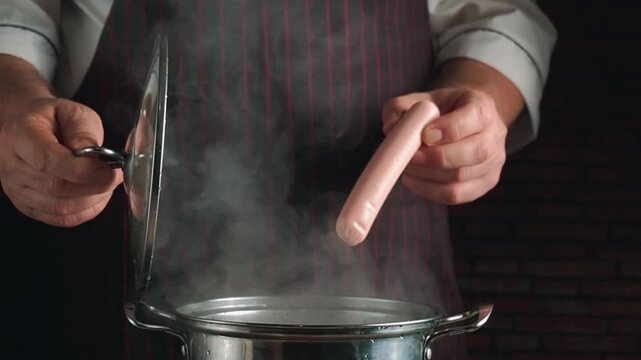 Chef in striped apron lifts pot lid, revealing steam and sausage while preparing meal in a cozy kitchen with brick wall background
