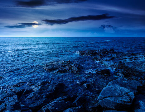 sea coast with stones and boulders at night. view of beach with rock in seaweed. full moon over dynamic cloudscape on the sky above horizon. mysterious summer vacations background concept