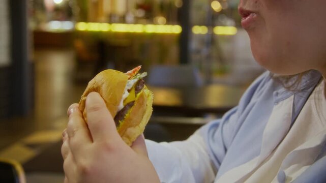 Close up shot of young woman taking bite of big burger with pleasure, holding it with both hands in modern fast food cafe