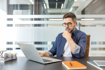 Naklejka premium Confident businessman in a modern office concentrates on his laptop, analyzing strategies and ideas for a complex project while planning solutions and next steps in his workday