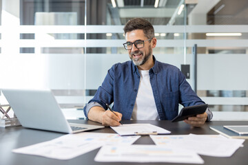 Obraz premium Smiling businessman working on a laptop, calculating finances with a calculator, and signing important paperwork in a modern office environment, representing success and financial management