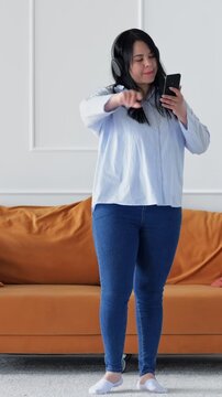 Caucasian Woman Standing Near Orange Couch Dancing With Headphones, Scrolling Phone Between Moves, Upbeat Music And Joyful Movement, Casual Jeans And Socks, Modern Living Interior, Carefree Energy