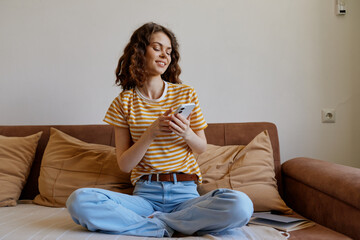 Fototapeta premium A relaxed woman sits crosslegged on a sofa at home, smiling while scrolling her phone. A casual lifestyle scene focused on comfort, quiet moments, and everyday connectivity.