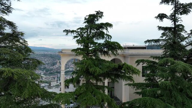 Aerial view of Mtatsminda Park funicular station and restaurant building in Tbilisi, Georgia, with the TV tower and Ferris wheel in a lush green mountain landscape.