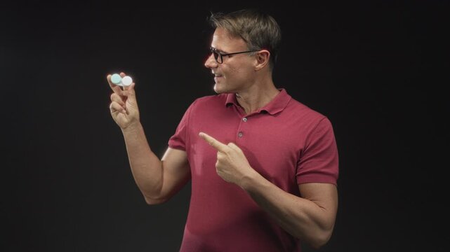 Man wearing glasses and red polo, holding a contact lens case between thumb and forefinger and points finger at it in studio; eye care confidence.