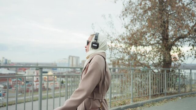 Caucasian woman walking by railing overlooking city skyline wearing trench coat handbag and headphones confident stride commuter content creator urban autumn scene subtle wind city view