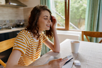 Young woman with curly hair sits at a sunlit kitchen table, smiling softly while tapping a tablet in a striped yellow shirt. © SHOTPRIME STUDIO
