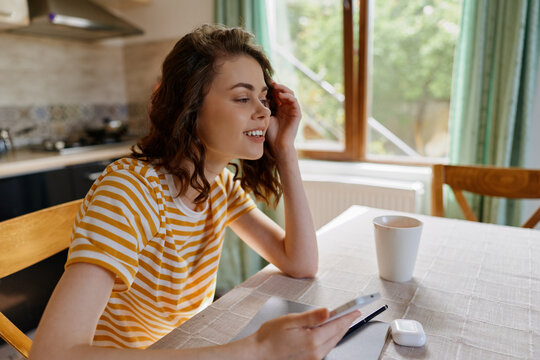 Young woman with curly hair sits at a sunlit kitchen table, smiling softly while tapping a tablet in a striped yellow shirt.