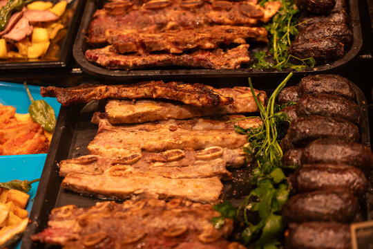 Trays of roasted pork ribs and dark blood sausages (morcella) garnished with parsley at a traditional food market.