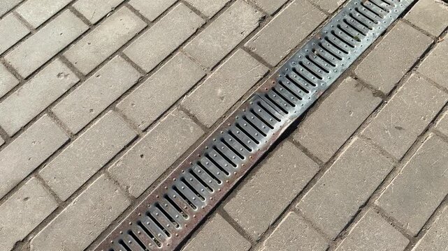 Metal grate spillway on paving stones white downpipe above, patterned metal cover set into paver joints, thin rust streaks