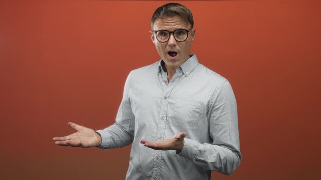 Man with open palms gesturing in studio, wearing light buttondown shirt and glasses, standing against orange backdrop, facing left; confusion questioning.