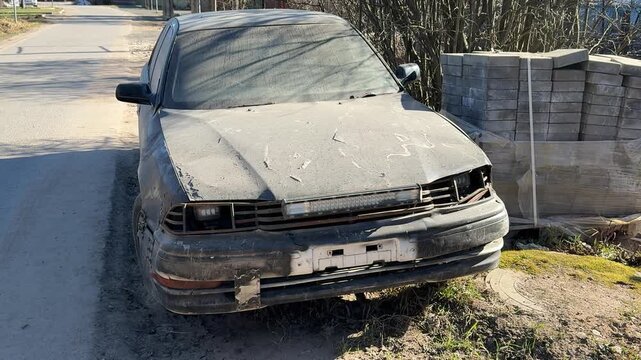 Dusty black sedan abandoned by roadside, frontend collision damage with cracked bumper, tinted windows covered in grime