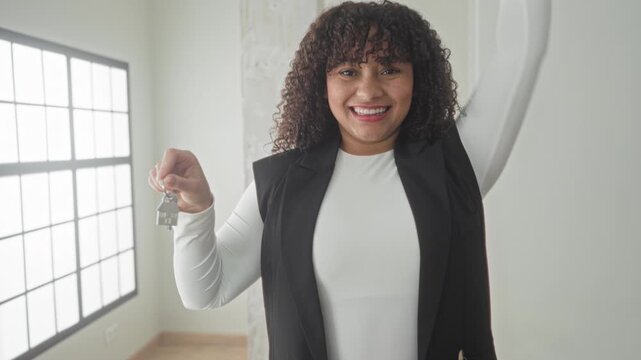 Hispanic woman holding house keys high, smiling with visible face and raised arm inside bright empty building; homeownership joy.