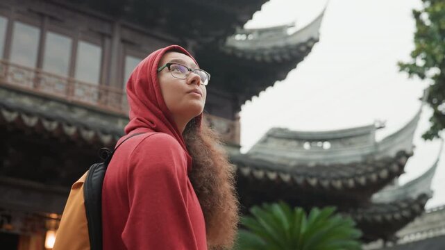 Solo traveler in red hoodie looking up at ornate pagoda roof and eaves, orange backpack visible glasses and curly hair, overcast sky, temple courtyard ambiance, sense of wonder and cultural discovery,