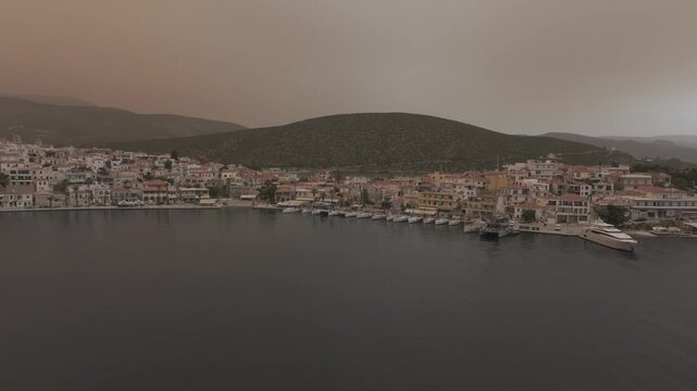 Cinematic drone flight approaching Ermioni harbor from the open sea revealing marina, coastal town and moody orange sky over Greece