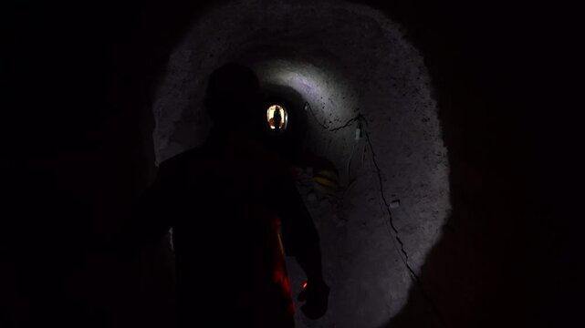 Cinematic silhouette tracking shot of a worker with a flashlight navigating a dark, rocky underground mine shaft. Dramatic, moody B-roll ideal for thrillers, mining docs, and rescue concepts.