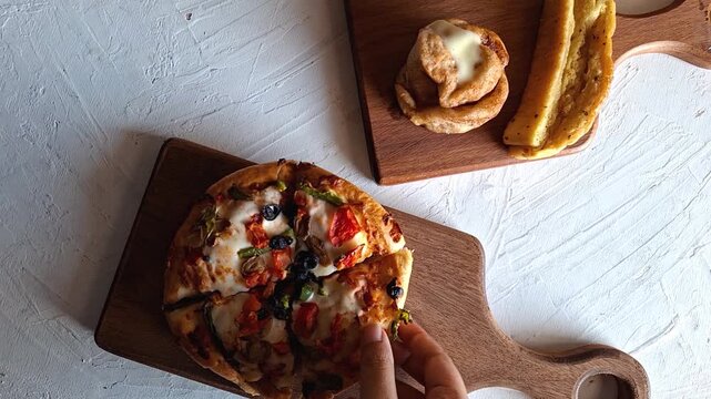 Small Homemade Veggie Pan-Style Pizza Served on Rustic Wooden Cutting Board with Cinnamon Roll and Slice of Breadstick on Textured White Surface.