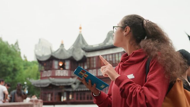 Young woman tourist reading guidebook near temple, red hoodie and backpack, pausing to study map and admire ornate pagoda architecture candid urban sightseeing moment with calm curiosity, heritage