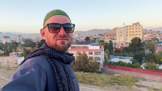 western tourist traveler visit kabul taking selfie at city viewpoint. man with sunglasses & shemagh scarf overlooking urban afghanistan landscape. travel vlogger, tourism, & exploration