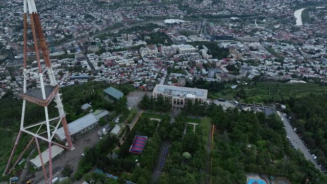 Aerial view of Mtatsminda Park funicular station and restaurant building in Tbilisi, Georgia, with the TV tower and Ferris wheel in a lush green mountain landscape.