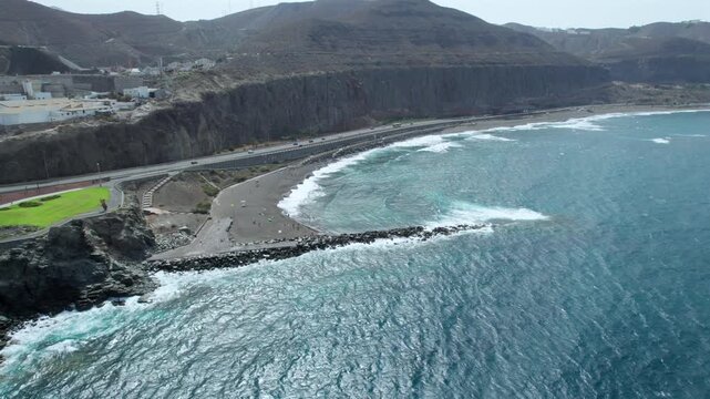 Stunning aerial perspective of La Laja beach in Las Palmas de Gran Canaria. Turquoise waves crash on the volcanic dark sand and rock jetty