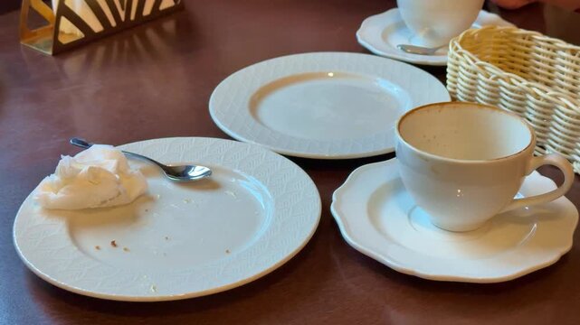 Empty coffee cup on saucer among empty plates and wicker basket, spoon with crumbs, close-up of ceramic tableware on wooden table
