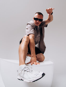 Young male hip hop dancer in sunglasses, shorts, and gray shirt posing on a gray studio background. Wide-angle low shot with dramatic shoe distortion.