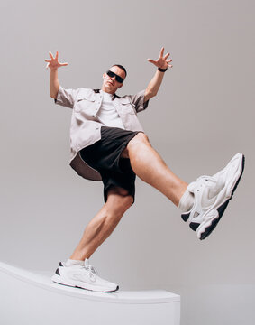 Young male hip hop dancer in sunglasses, shorts, and gray shirt posing on a gray studio background. Wide-angle low shot with dramatic shoe distortion.