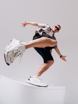 Young male hip hop dancer in sunglasses, shorts, and gray shirt posing on a gray studio background. Wide-angle low shot with dramatic shoe distortion.