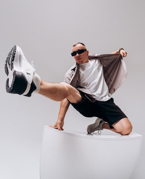 Young male hip hop dancer in sunglasses, shorts, and gray shirt posing on a gray studio background. Wide-angle low shot with dramatic shoe distortion.