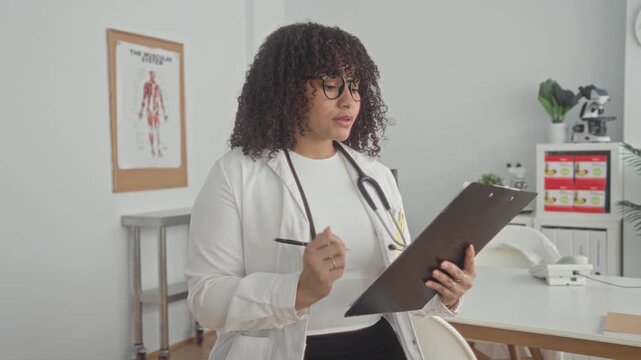Woman doctor writing on clipboard with stethoscope and glasses, hand holding pen in clinic building while checking notes; focused care.
