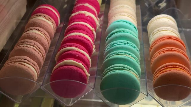 Colorful macarons coffee shop window close-up, rows of teal pink orange and brown shells arranged on clear tray inside warm lit
