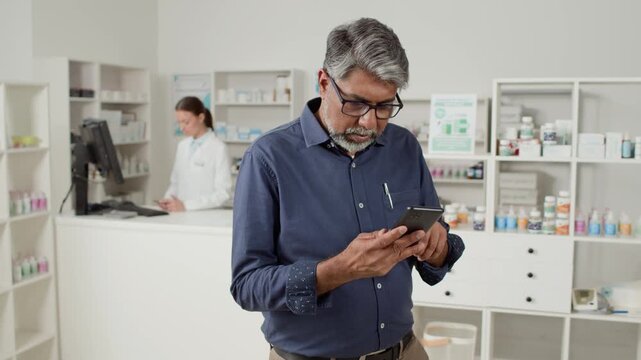 Medium shot of mature Indian man as male customer choosing medication or multivitamins at pharmacy and reading labels carefully using online medical advice via phone, copy space