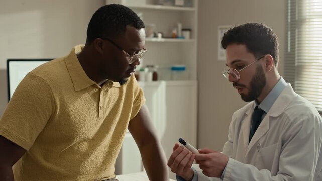 Side view waist up shot of Middle Eastern male doctor holding insulin pen in hands explaining its use to Black male patient with diabetes during medical appointment in modern clinic office