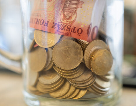 Clear jar holds Hungarian coins and banknotes. Forint coins and cash stack inside transparent container. Banknote rests atop coins. Image conveys savings or financial accumulation. Background softly b