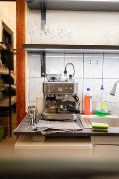 Modern espresso machine sits on kitchen counter. Silver and stainless steel finish, professional design. Glassware hangs above, tidy and organized. Sponge and cleaning supplies nearby, ready for use